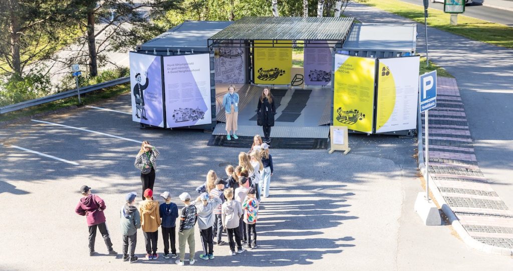 Eine Gruppe junge Menschen stehen auf eine Parkplatz vor einer in einem Container gestaltete Ausstellung